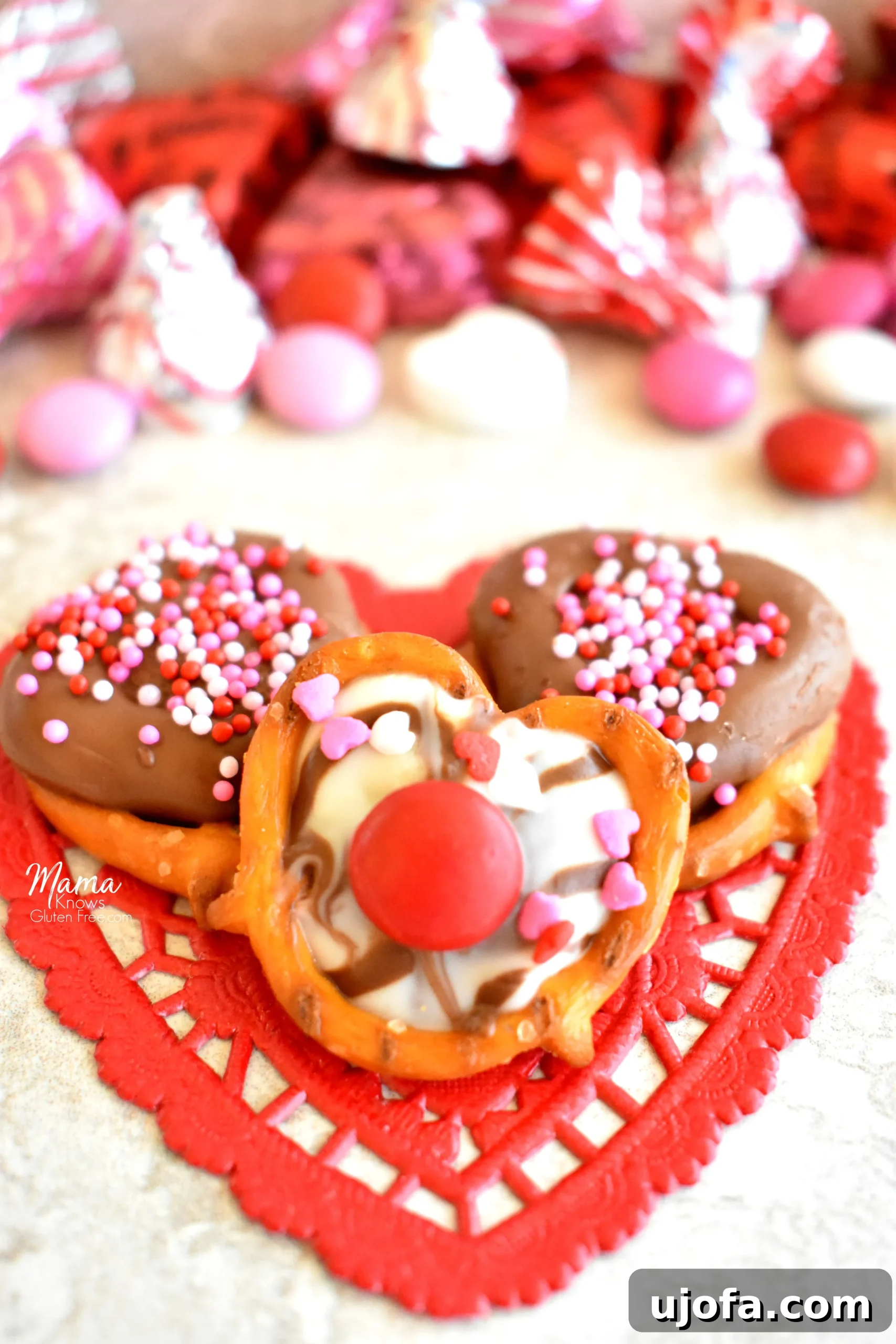 Irresistible Gluten Free Chocolate Pretzels for Valentines 4 Three beautifully decorated gluten-free chocolate covered pretzels resting on a red heart doily, with assorted Valentine's candies in the background
