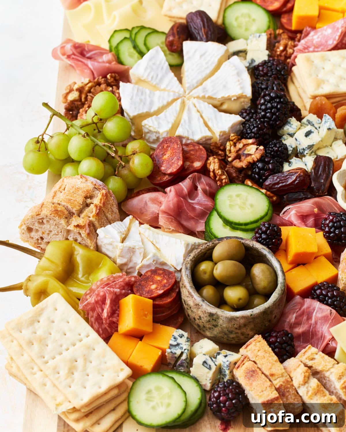 Gourmet Gluten-Free Charcuterie 5 A charcuterie board with Brie cheese wedges at the center surrounded by meats like salami and prosciutto, fresh cucumber slices, green grapes, cheddar cubes, and assorted crackers. A small bowl of green olives is placed on the board.