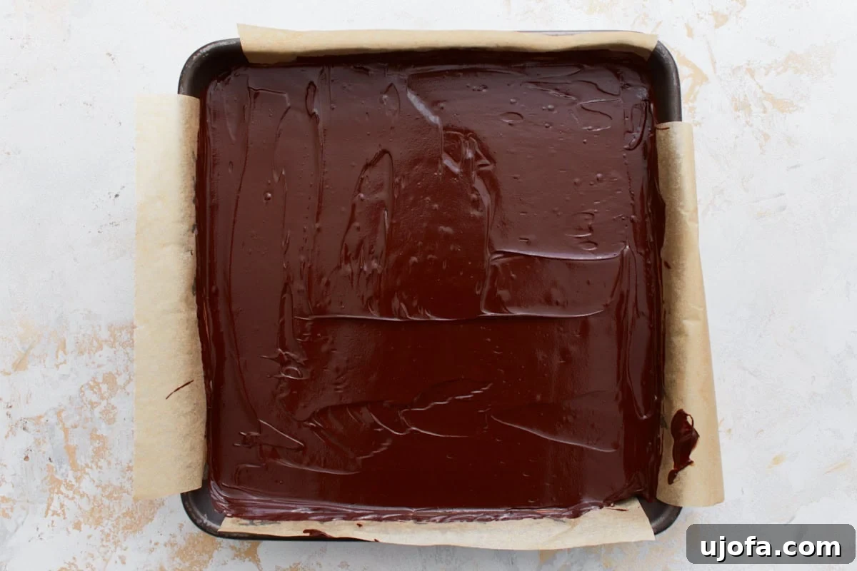 A parchment-lined baking pan with a layer of smooth melted chocolate spread over the caramel, completing the top layer.