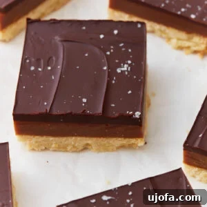 Close-up of a slice of gluten-free millionaire shortbread topped with glossy chocolate, a layer of caramel, and a shortbread base, sprinkled with flaky sea salt.