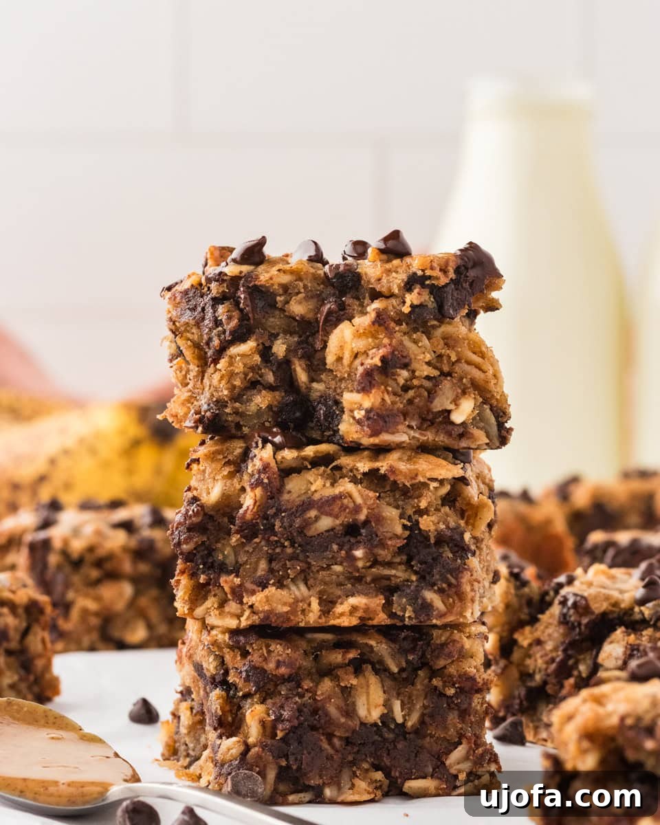Golden Banana Oat Bars 2 A stack of three soft-baked banana oatmeal bars with chocolate chips, showcasing their chewy texture. A bottle of milk and ripe bananas are blurred in the background.