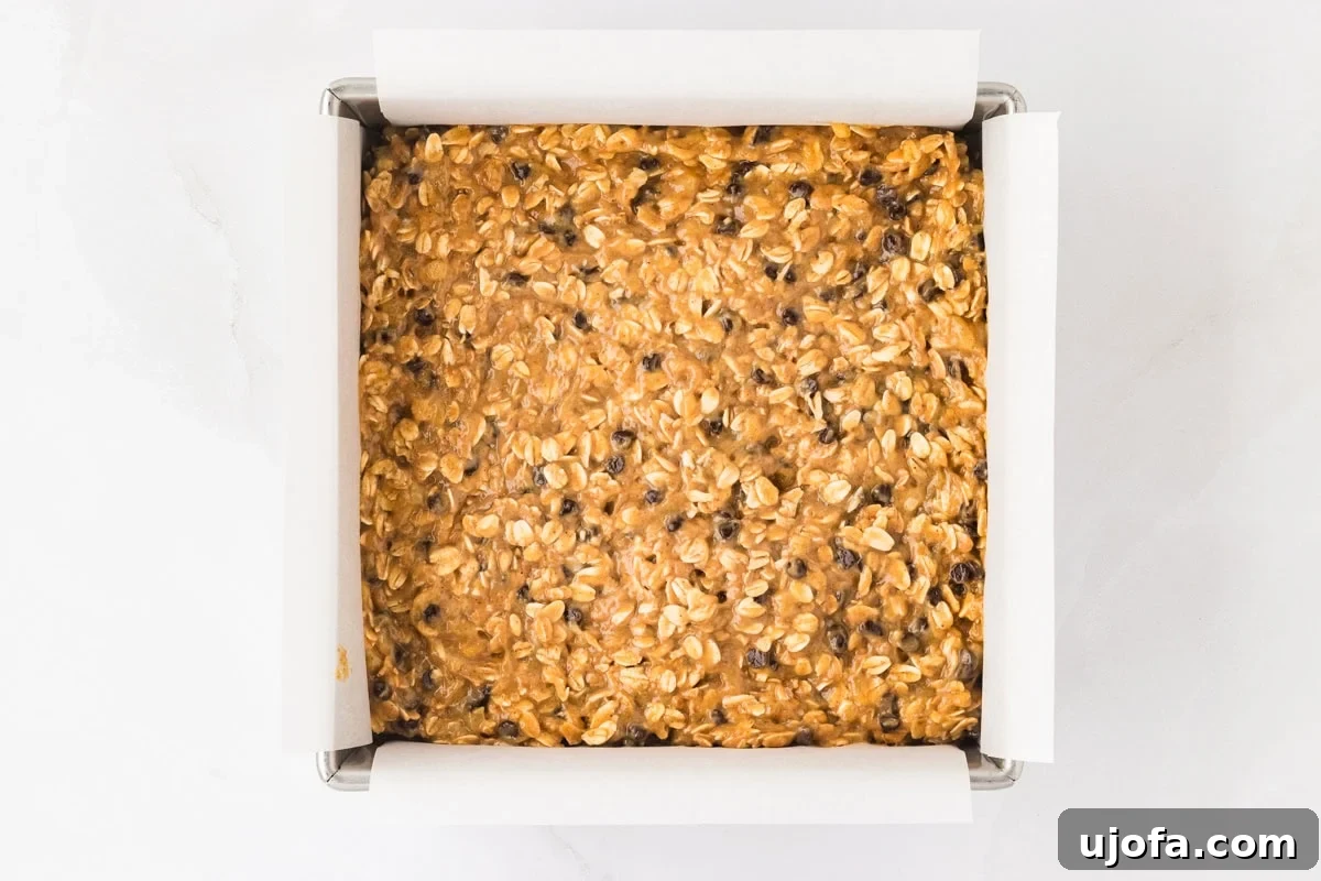 Golden Banana Oat Bars 11 A square baking pan lined with parchment paper, filled with unbaked banana oatmeal bar batter, showcasing visible oats and chocolate chips.