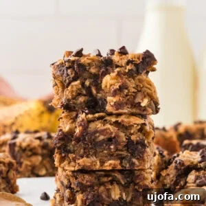 A stack of three soft-baked banana oatmeal bars with chocolate chips, showcasing their chewy texture. A bottle of milk and ripe bananas are blurred in the background.