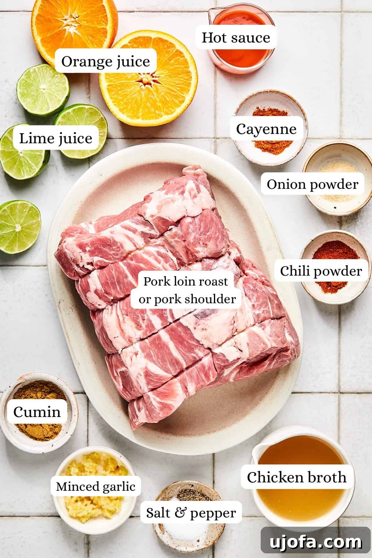 Overhead view of fresh ingredients for pork carnitas: pork roast, aromatic spices, whole garlic cloves, citrus fruits (lime and orange), and a measuring cup of chicken broth
