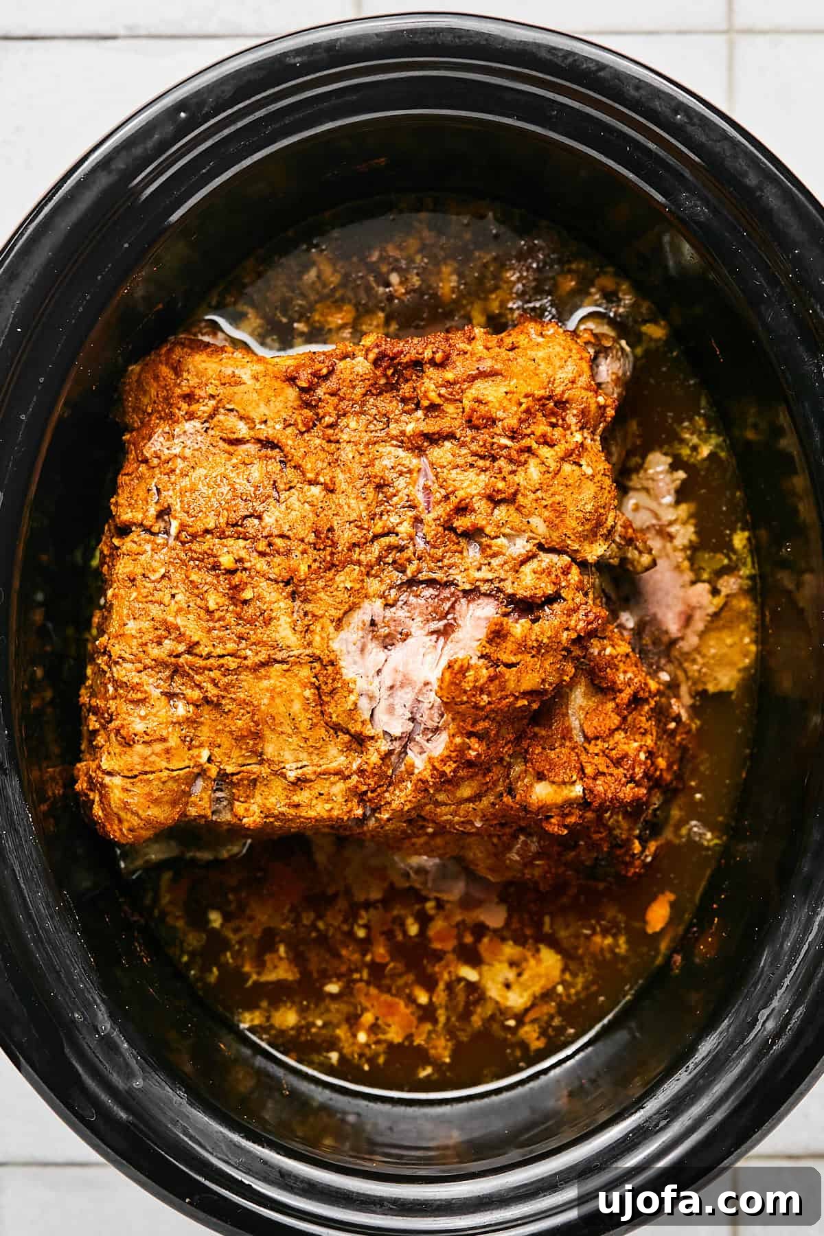 Pork roast immersed in a rich liquid within the slow cooker, surrounded by minced garlic, slices of lime and orange, and chicken broth, simmering gently