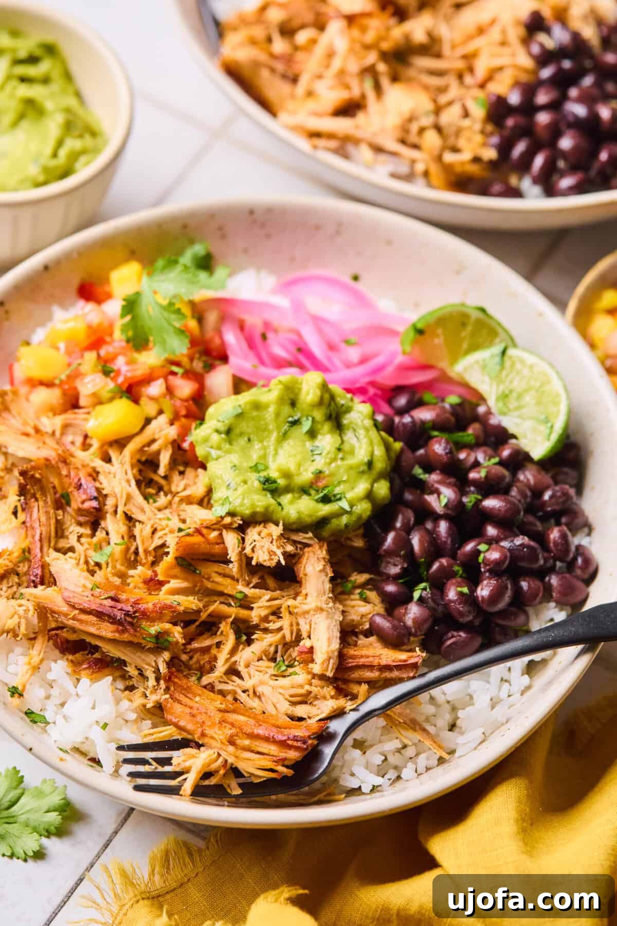 A vibrant bowl of slow cooker carnitas, artfully arranged with creamy guacamole, savory black beans, fluffy cilantro lime rice, tangy pickled onions, and a colorful mango salsa