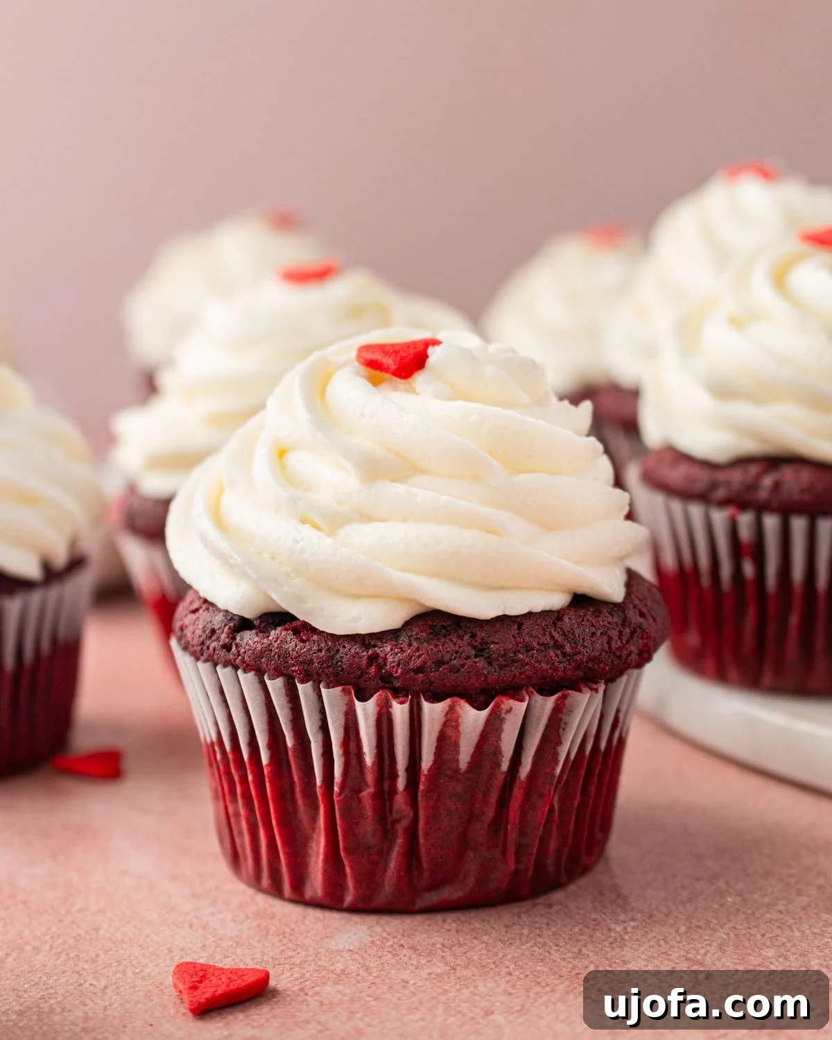 Close-up of a gluten-free red velvet cupcake with creamy swirled cream cheese frosting, topped with a small red heart decoration. Several cupcakes are in the background, slightly blurred.