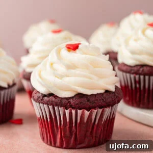 Close-up of a gluten-free red velvet cupcake with creamy swirled cream cheese frosting, topped with a small red heart decoration. Several cupcakes are in the background, slightly blurred.