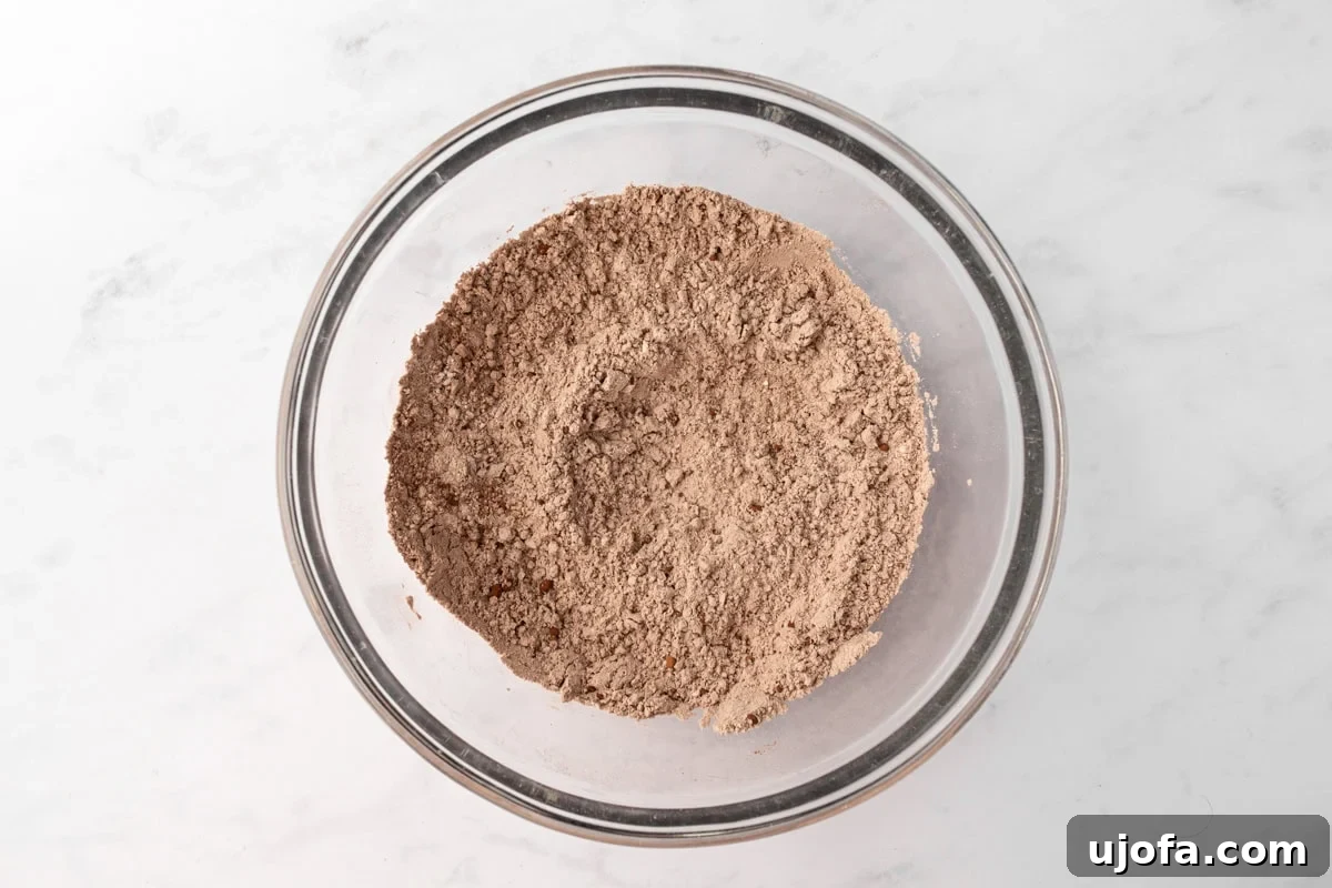 A glass mixing bowl filled with dry ingredients, including cocoa powder and gluten-free flour, sitting on a white marble surface.
