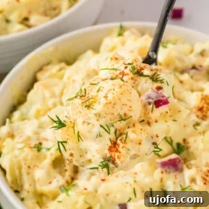 A close-up of a spoonful of creamy gluten-free potato salad with diced potatoes, red onion, and fresh dill, sprinkled with paprika.