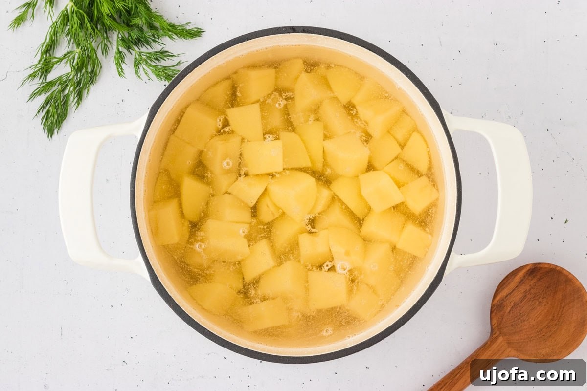 A pot of boiling cubed potatoes on a white countertop, with fresh dill in the background, preparing for a gluten-free potato salad.