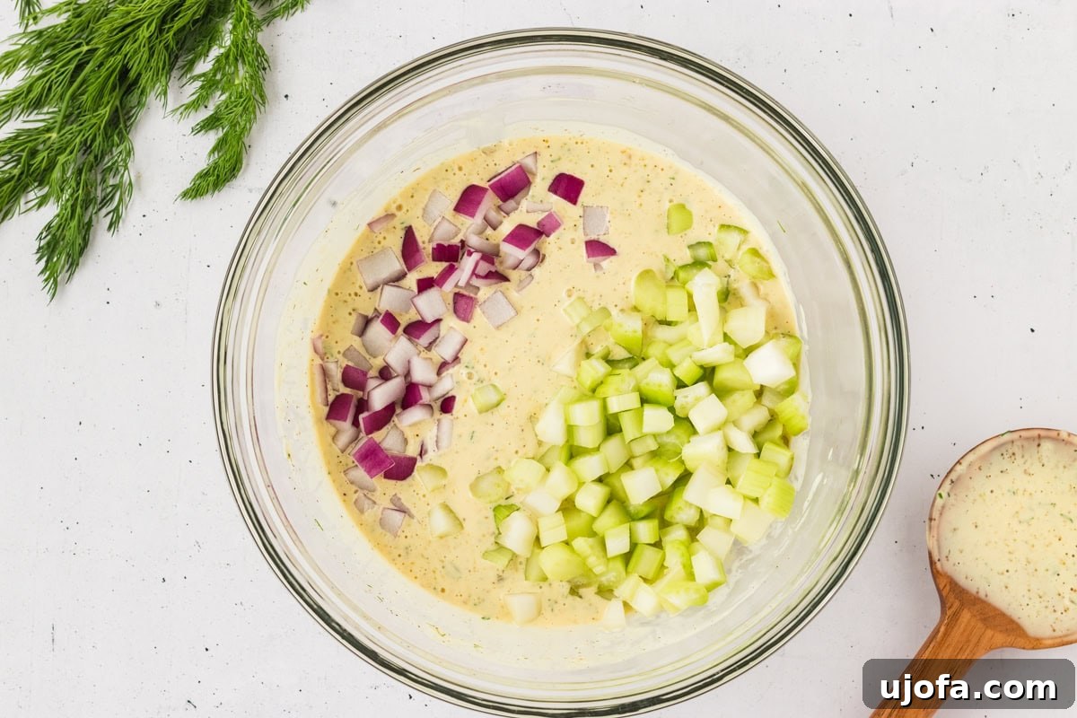 A glass bowl with gluten-free potato salad dressing, mixed with diced red onion and chopped celery, ready for combining with potatoes.