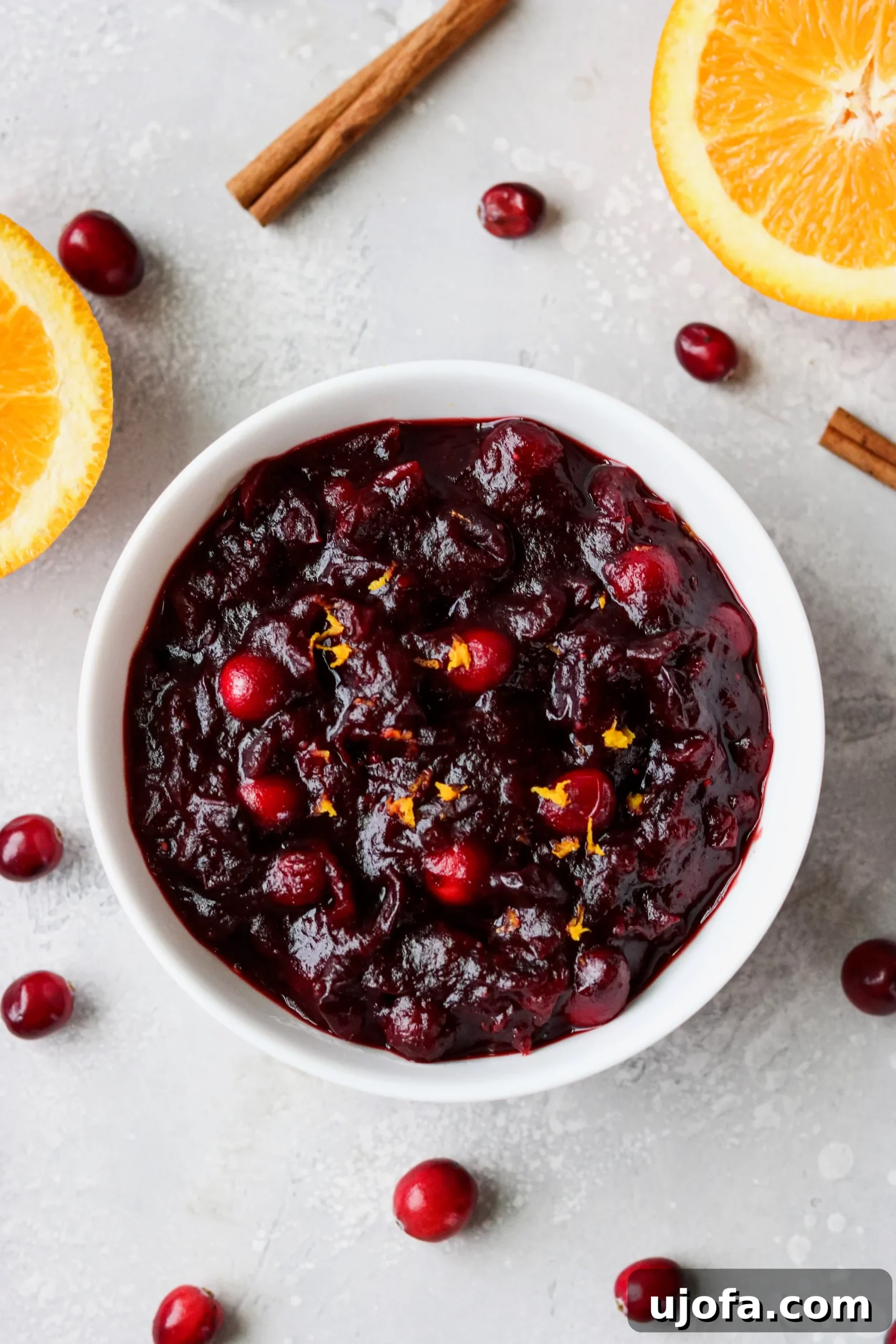Muhteşem Paleo Şükran Günü Sofrası İçin Her Şey 9 A vibrant, glossy cranberry sauce in a white bowl, garnished with fresh orange slices and rosemary sprigs, showcasing its rich red color and inviting texture.