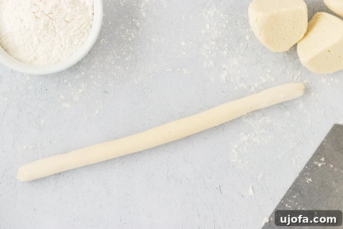 A perfectly rolled, elongated log of cauliflower gnocchi dough, consistently about three-quarters of an inch thick, resting on a lightly floured surface. Nearby are additional portions of the dough and a bowl of flour, ready to continue the shaping process.
