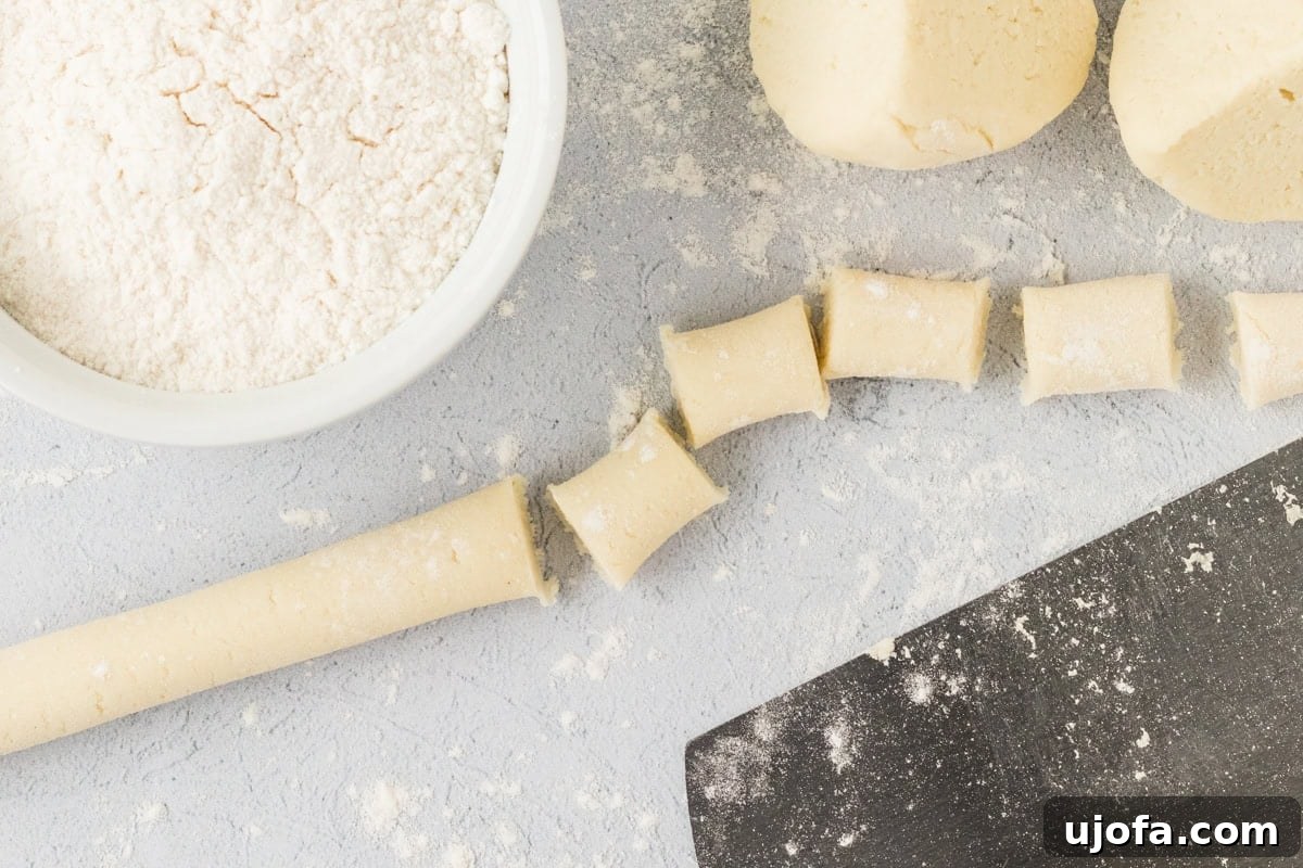 A close-up view of a rolled log of cauliflower gnocchi dough, meticulously cut into numerous small, bite-sized, 1-inch pieces. These pieces are laid out on a flour-dusted surface, with a bowl of flour and a dough scraper nearby, indicating readiness for the next shaping stage or cooking.