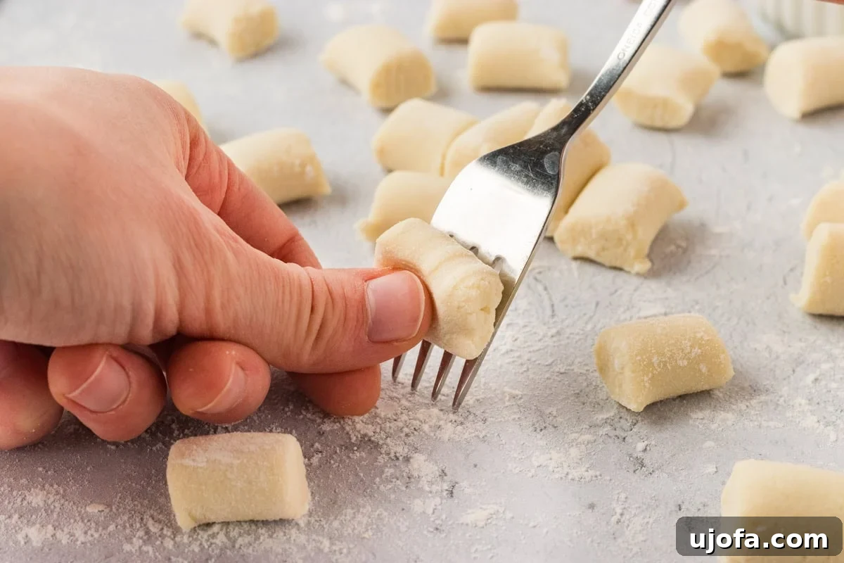 A skilled hand gently pressing a small, rectangular piece of cauliflower gnocchi dough against the tines of a fork, carefully creating distinct, parallel ridges on its surface. Several other perfectly formed gnocchi pieces are visible on a lightly floured surface, ready for cooking.