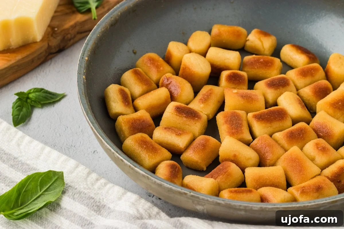 A vibrant close-up of golden brown, perfectly pan-fried cauliflower gnocchi sizzling enticingly in a well-seasoned skillet. Fresh basil leaves are artfully scattered amongst the gnocchi, adding a pop of color and aroma. A wedge of Parmesan cheese sits nearby, hinting at a delicious, ready-to-serve meal.