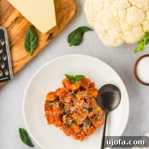 A white plate filled with golden brown cauliflower gnocchi covered in tomato sauce, garnished with fresh basil and grated cheese. A black spoon rests on the plate, surrounded by ingredients like Parmesan, fresh basil, and a head of cauliflower.