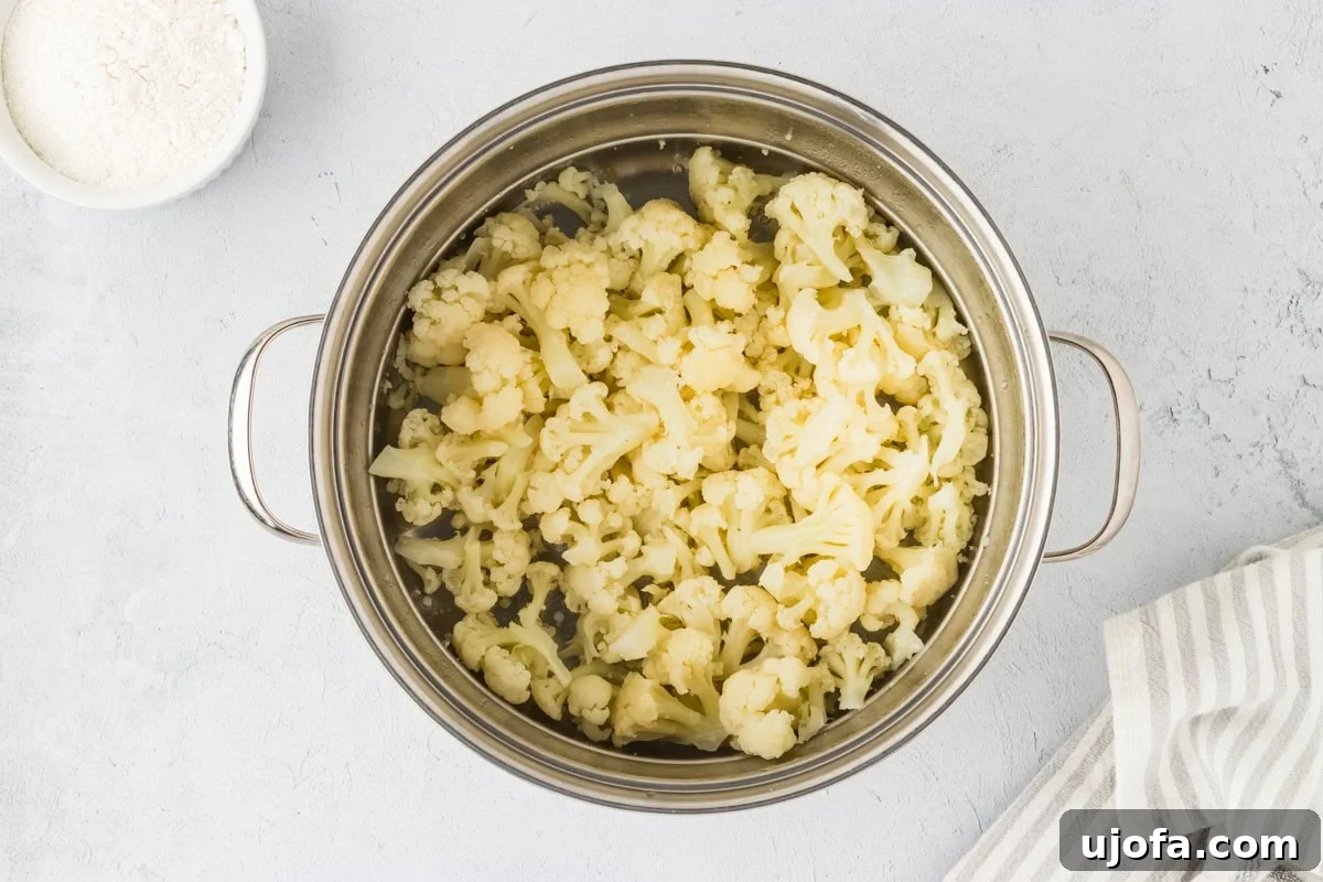A large cooking pot filled with freshly steamed, tender cauliflower florets. The florets are glistening with moisture and appear soft, indicating they are perfectly cooked and ready to be mashed and used in the gnocchi dough.