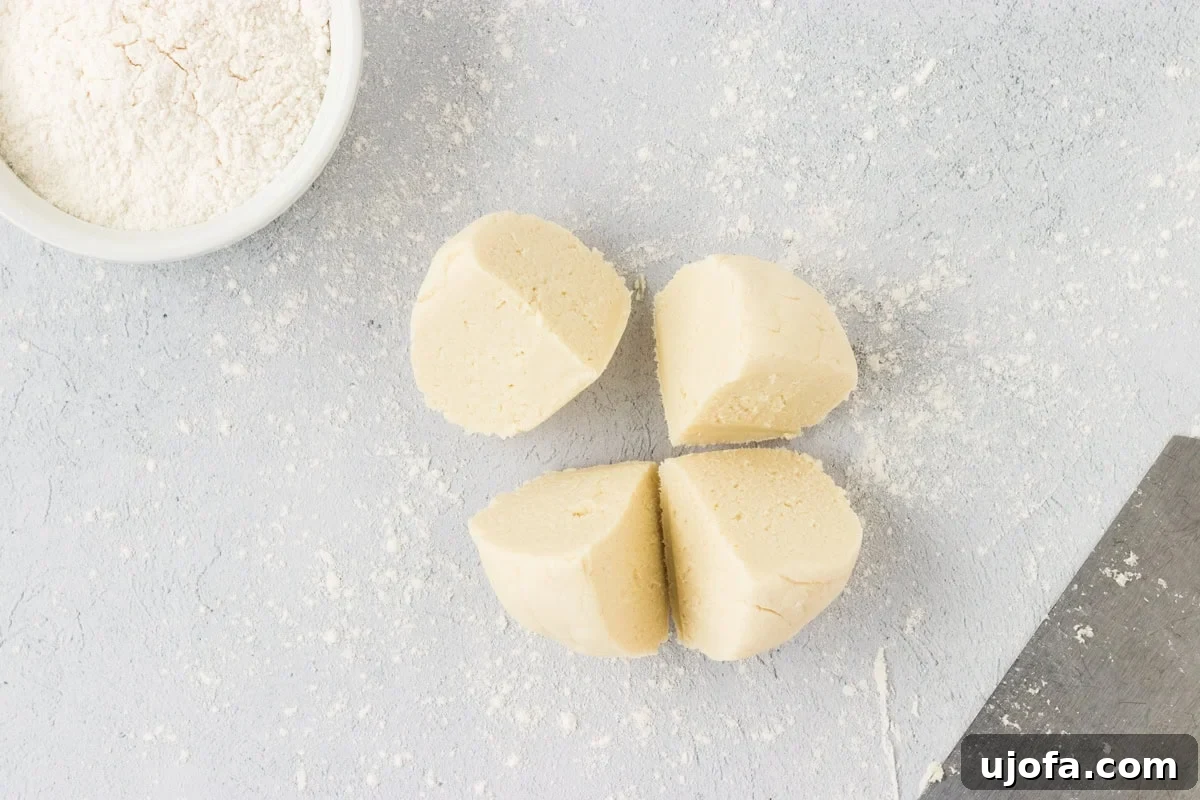A round ball of cauliflower gnocchi dough precisely divided into four equal portions using a metal dough scraper. The portions are neatly arranged on a lightly floured countertop, indicating readiness for the next step of rolling them into logs.