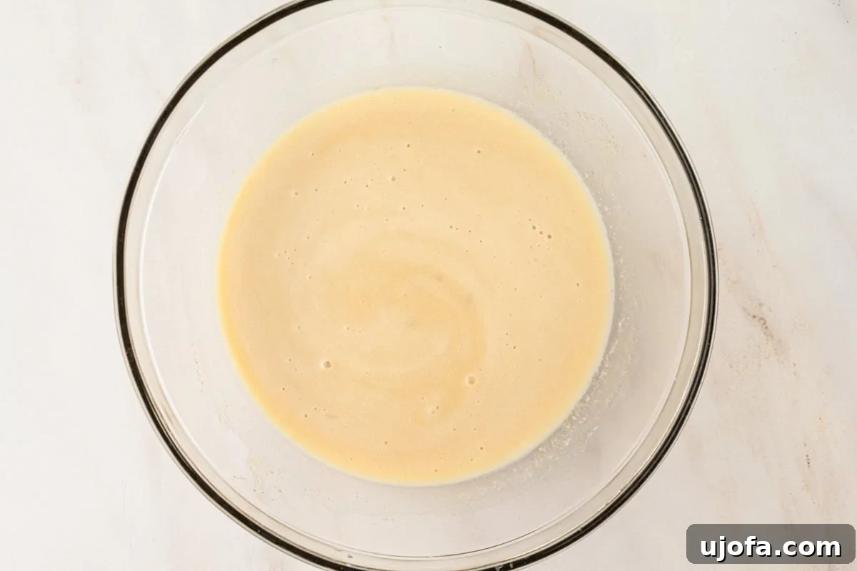 A glass mixing bowl filled with combined wet ingredients for the donut batter, showing a smooth and slightly frothy texture.