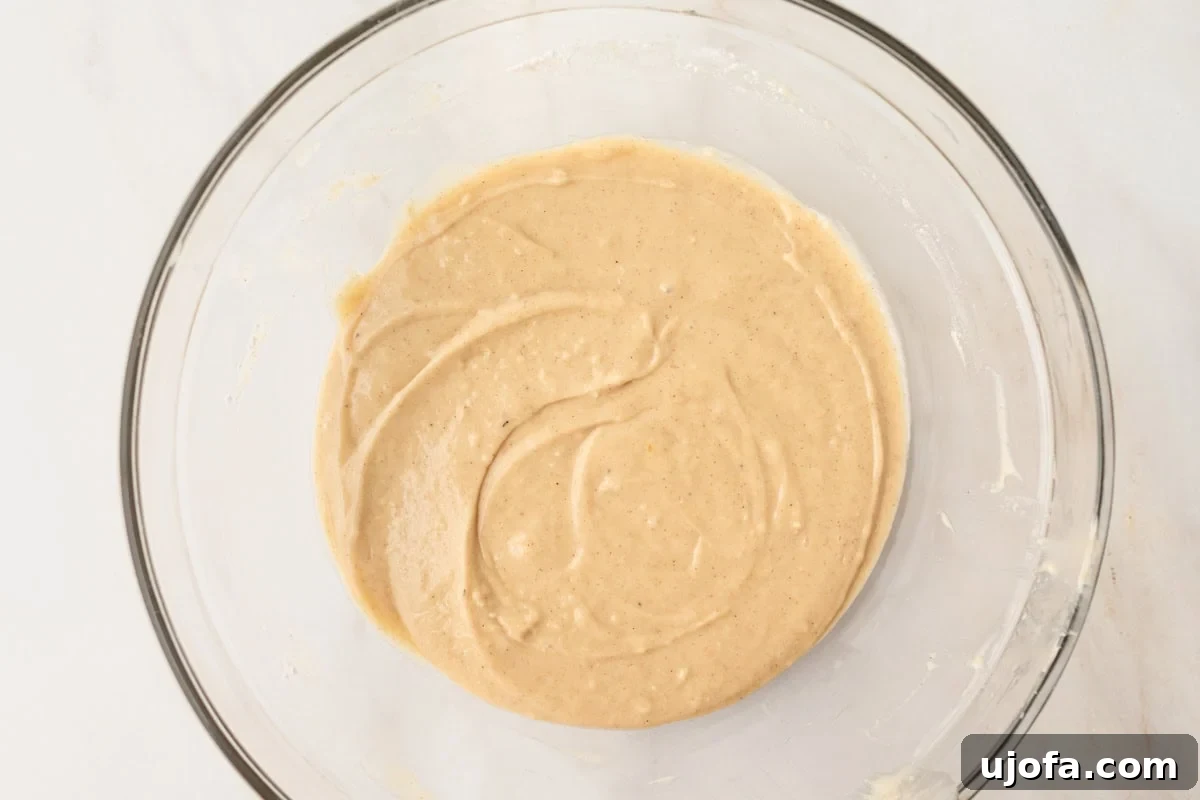A glass mixing bowl containing fully mixed gluten-free donut batter with a slightly speckled appearance from cinnamon and nutmeg.