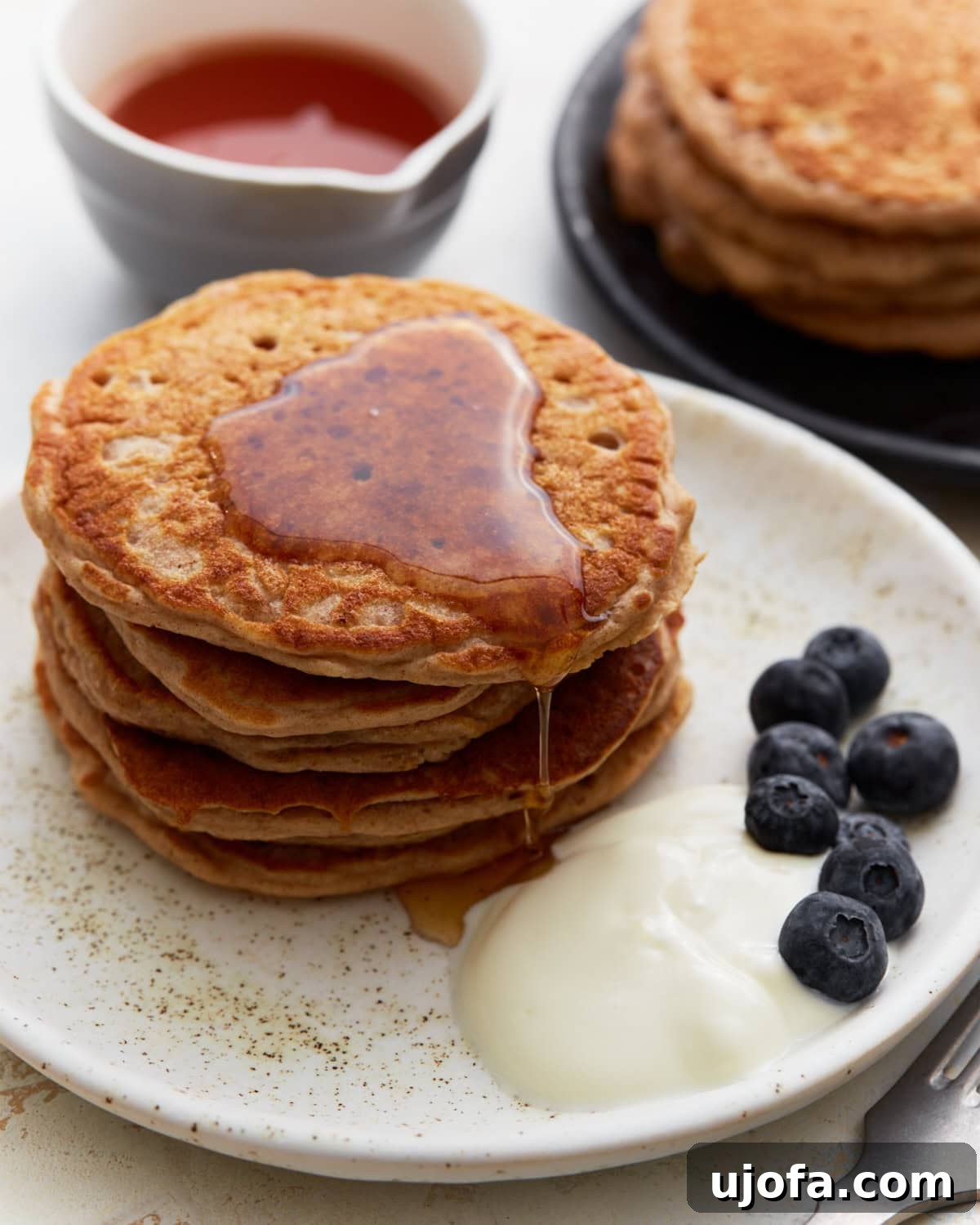 Hearty Buckwheat Flapjacks 2 A stack of golden-brown buckwheat pancakes on a white plate, drizzled with maple syrup, served with a side of yogurt and fresh blueberries. A small bowl of syrup and another plate of pancakes are in the background.