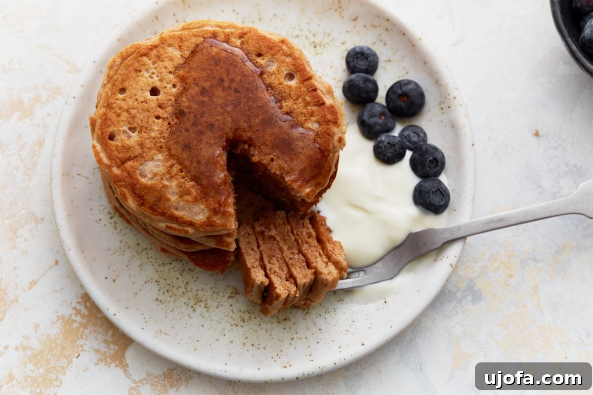 Hearty Buckwheat Flapjacks 10 A close-up of a stack of buckwheat pancakes with a cut-out section, revealing their soft, fluffy texture. The plate is garnished with fresh blueberries, yogurt, and a drizzle of maple syrup.