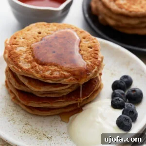 A stack of golden-brown buckwheat pancakes on a white plate, drizzled with maple syrup, served with a side of yogurt and fresh blueberries. A small bowl of syrup and another plate of pancakes are in the background.