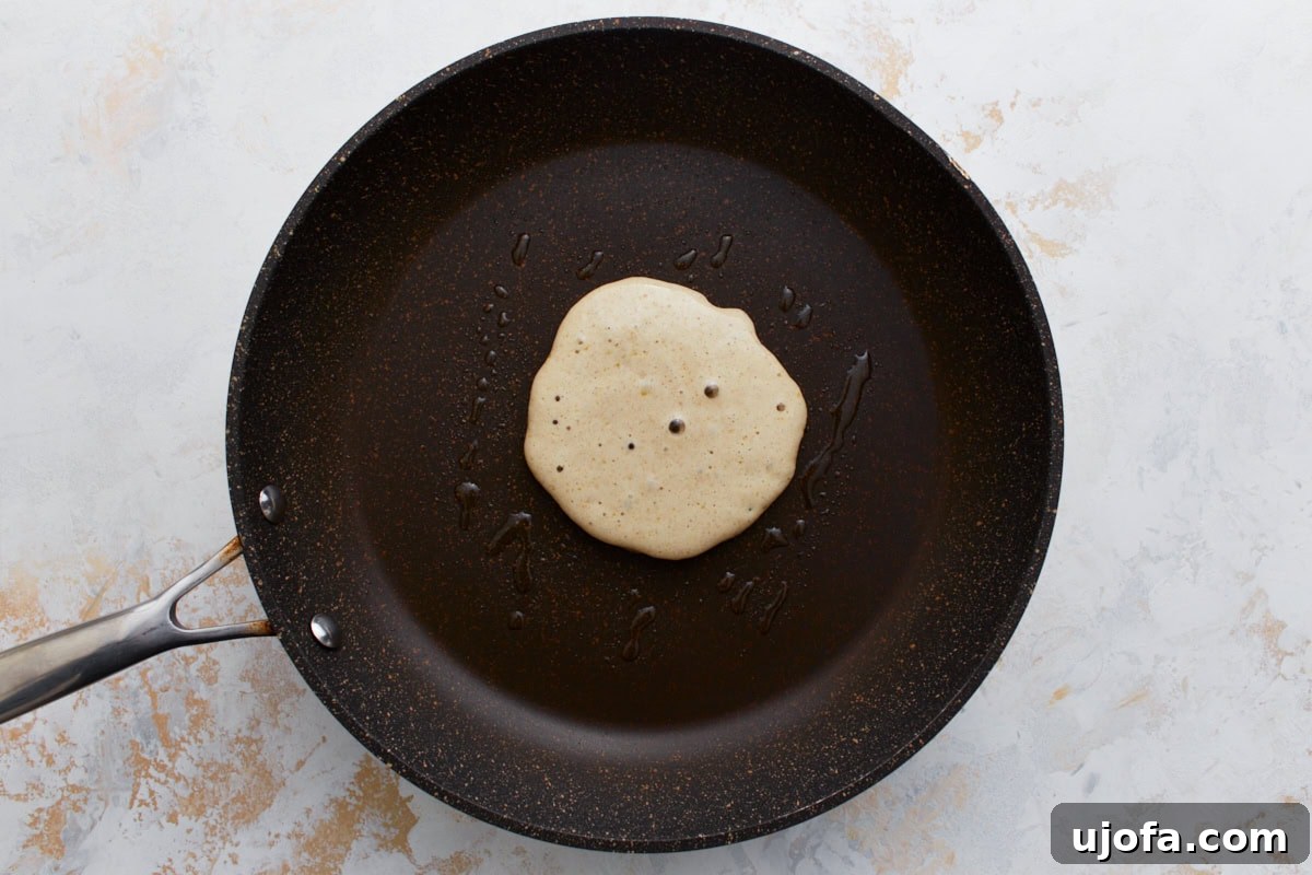 Hearty Buckwheat Flapjacks 8 A single buckwheat pancake cooking in a lightly greased non-stick pan, with small bubbles forming on the surface, showing the early stage of cooking.