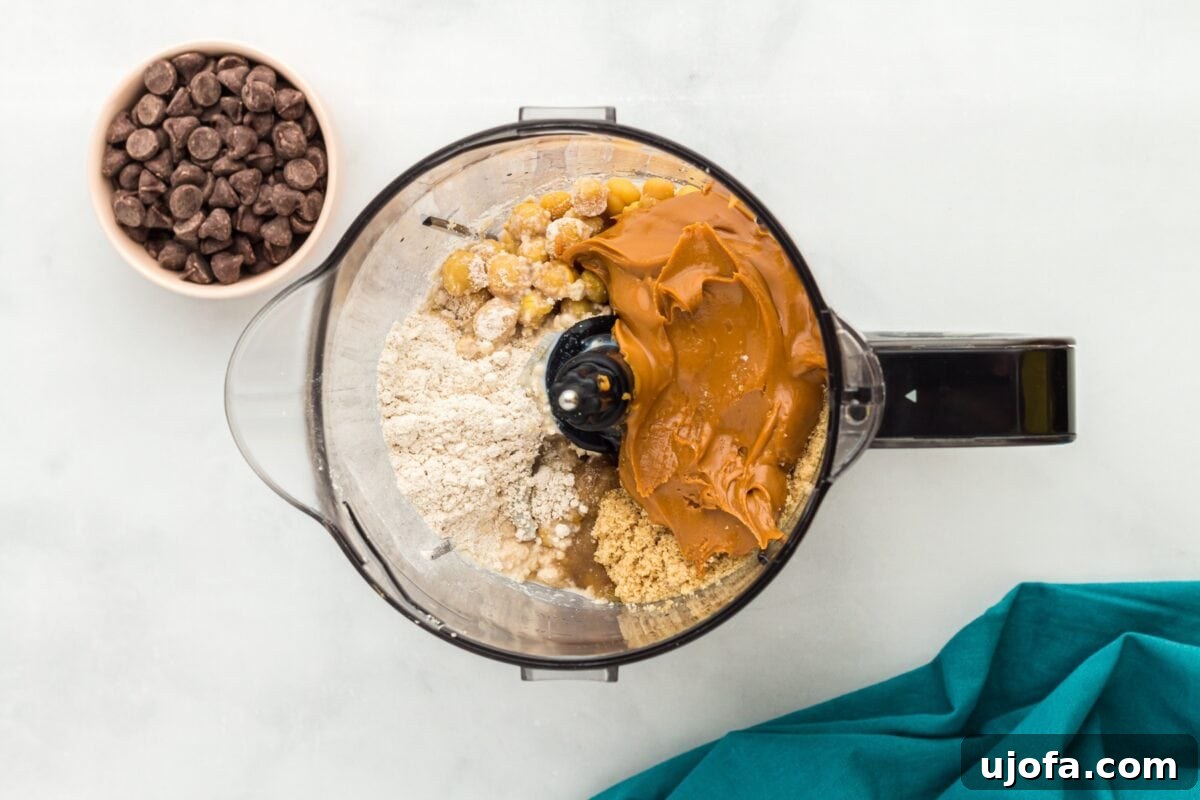 A top-down view of a food processor containing chickpeas, oat flour, peanut butter, and other ingredients, with a bowl of chocolate chips beside it.