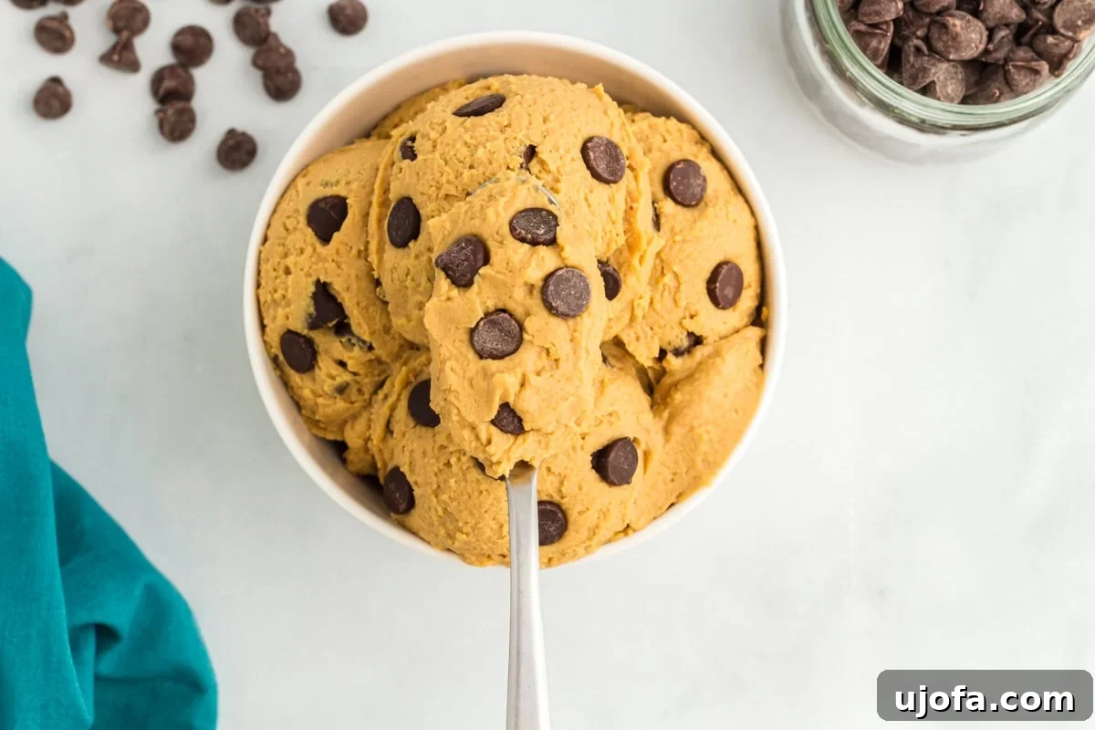 A white bowl filled with scoops of edible chickpea cookie dough, with chocolate chips sprinkled throughout. A spoon is inserted into the bowl.