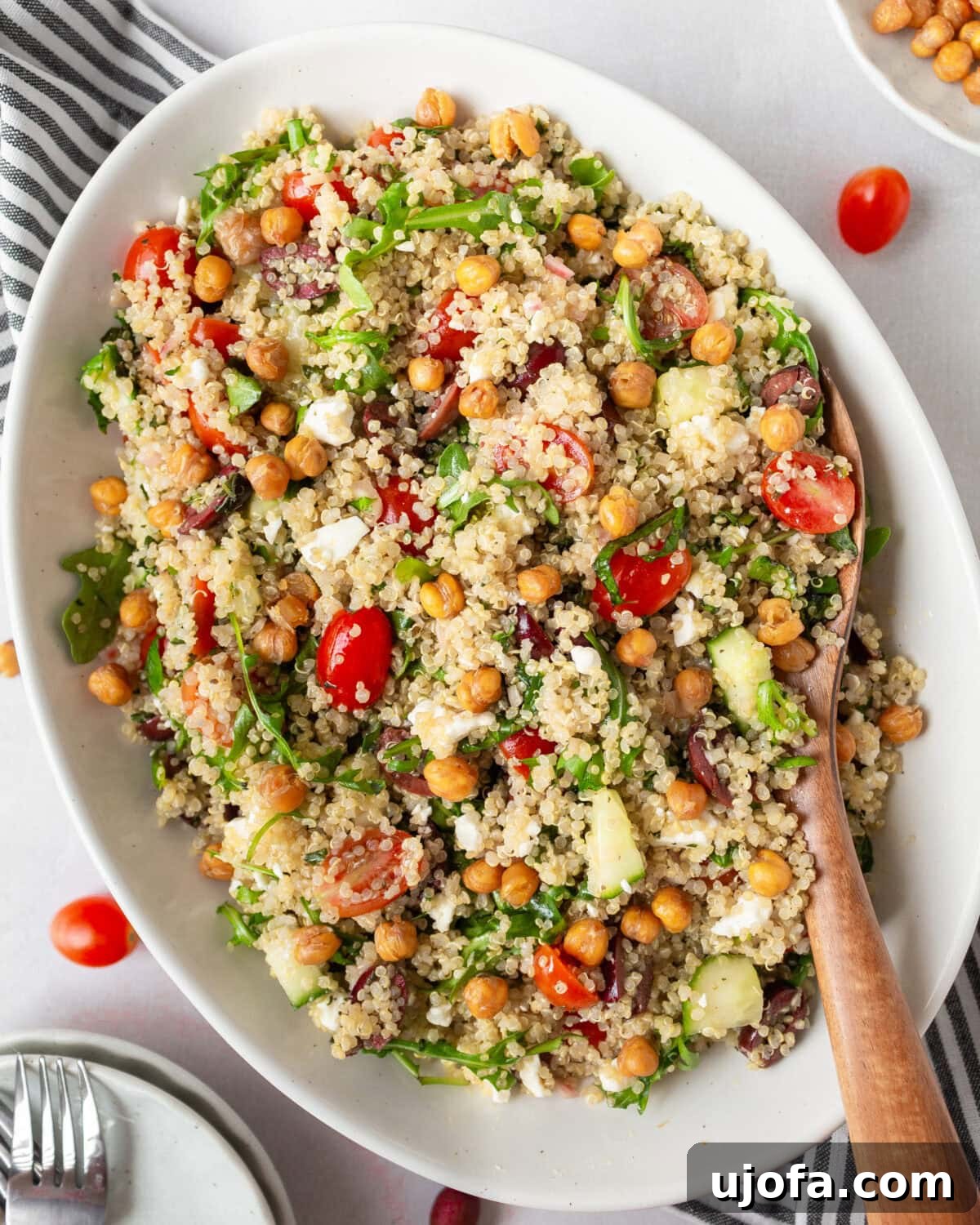 A white oval dish filled with Greek quinoa salad, featuring fluffy quinoa, cherry tomatoes, cucumbers, Kalamata olives, arugula, feta cheese, and crispy roasted chickpeas. A wooden spoon rests in the dish.