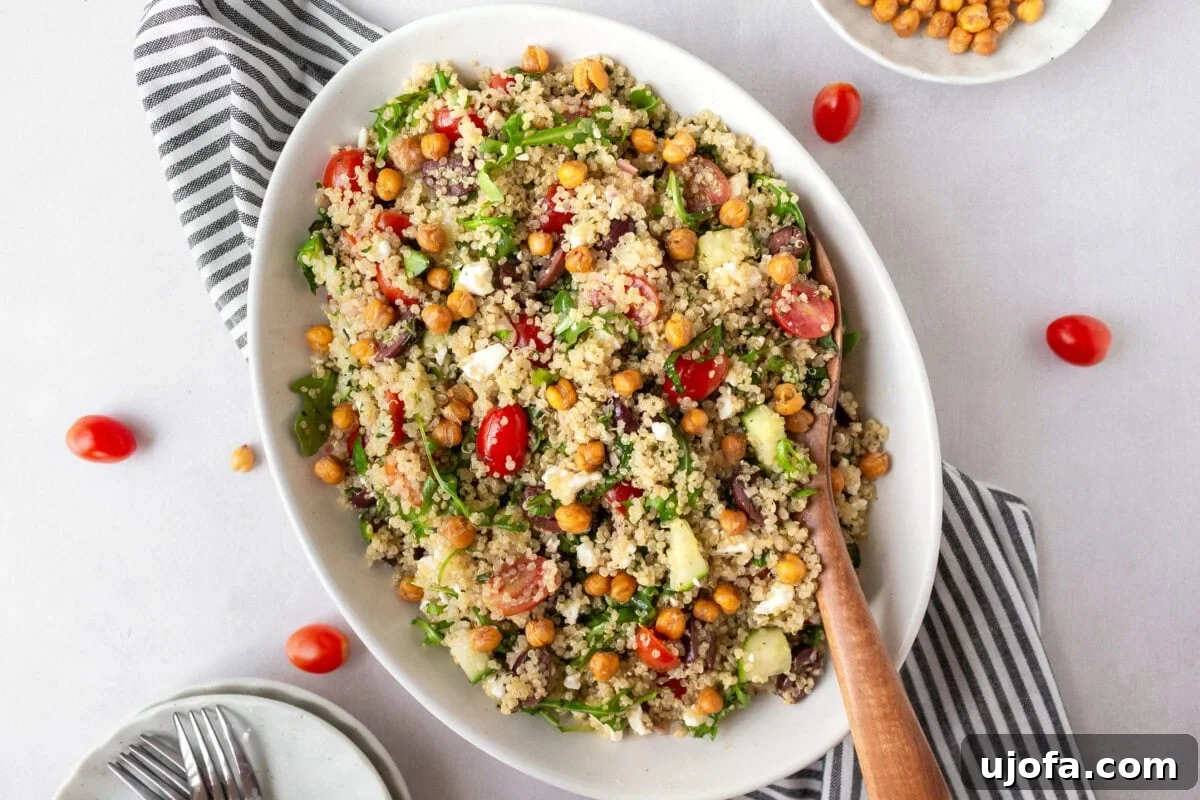 A fully assembled Greek quinoa salad served in a white oval dish, garnished with crispy roasted chickpeas and fresh herbs. A striped napkin and extra tomatoes surround the dish.