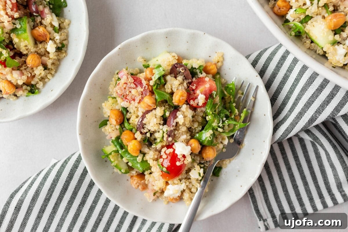 Small white bowls filled with Greek quinoa salad, ready to be served, with a striped napkin underneath and a fork resting in one bowl.