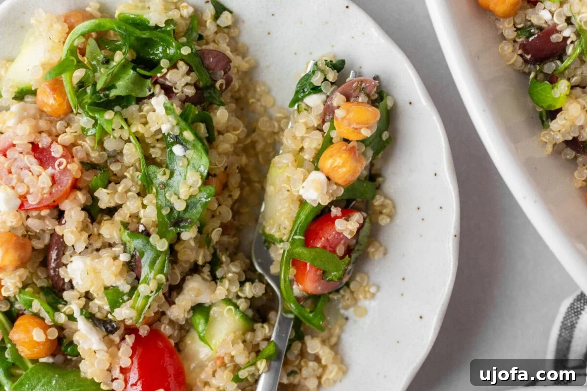 A close-up shot of a forkful of Greek quinoa salad, showcasing fluffy quinoa, arugula, cherry tomatoes, Kalamata olives, crispy roasted chickpeas, cucumbers, and crumbles of feta cheese on a white speckled plate. The vibrant ingredients highlight the fresh and flavorful textures of the dish.