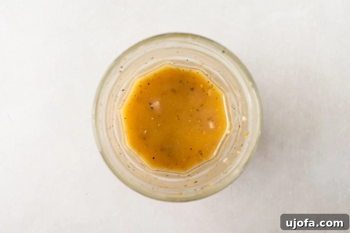 A close-up top-down view of a glass jar containing homemade Greek dressing, a golden mixture speckled with herbs and small bits of shallots or garlic.