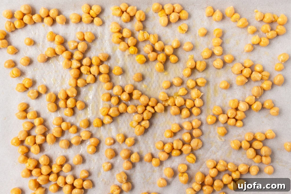A parchment-lined baking sheet with chickpeas coated in olive oil, evenly spread out before roasting.