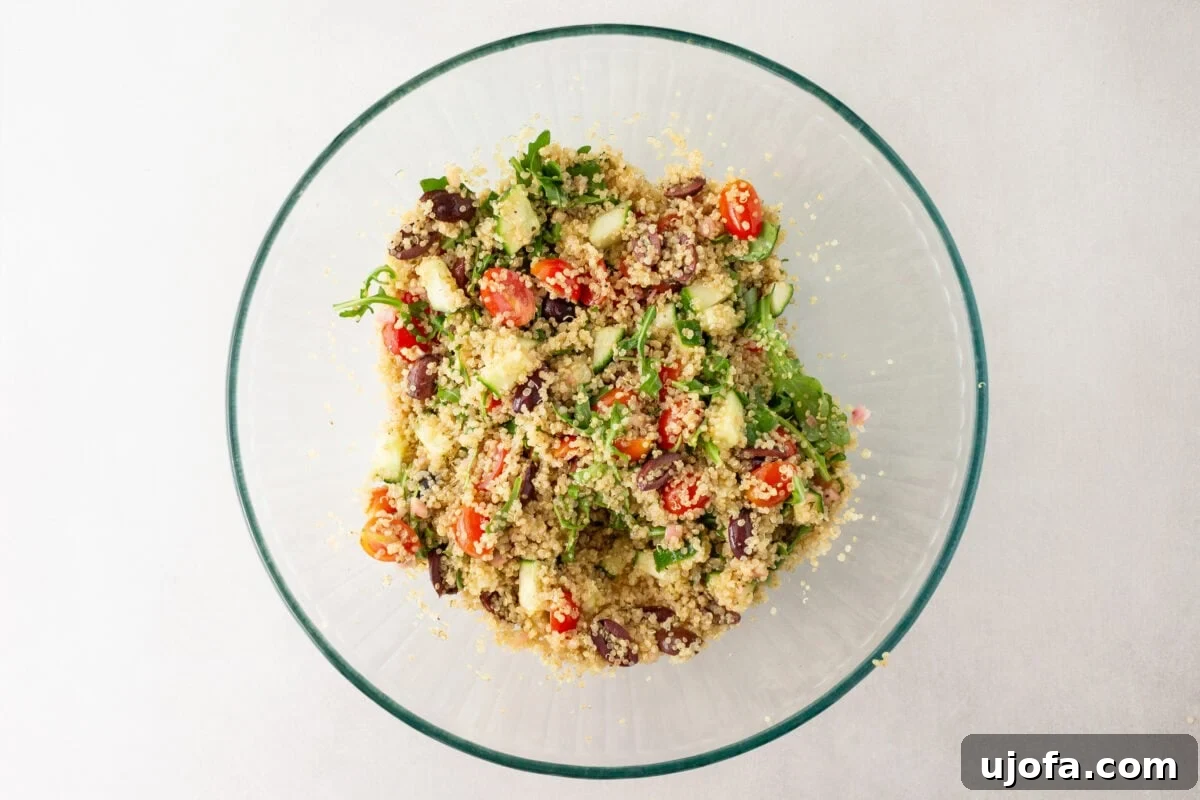 A glass bowl filled with freshly mixed Greek quinoa salad, showcasing bright ingredients like cherry tomatoes, cucumbers, Kalamata olives, arugula, and feta cheese.