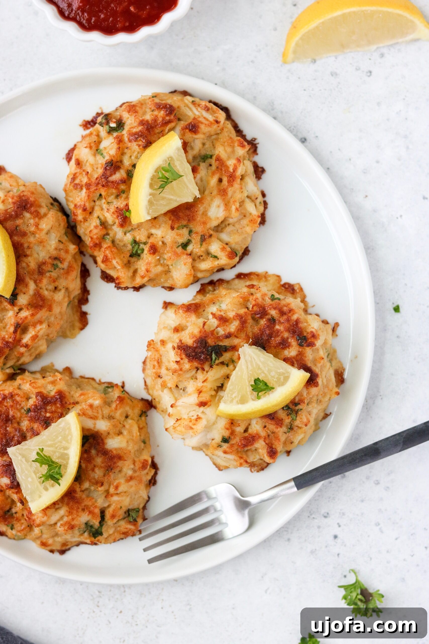 Four golden-brown keto crab cakes presented on a pristine white plate, accompanied by a silver fork, highlighting their exquisite texture.