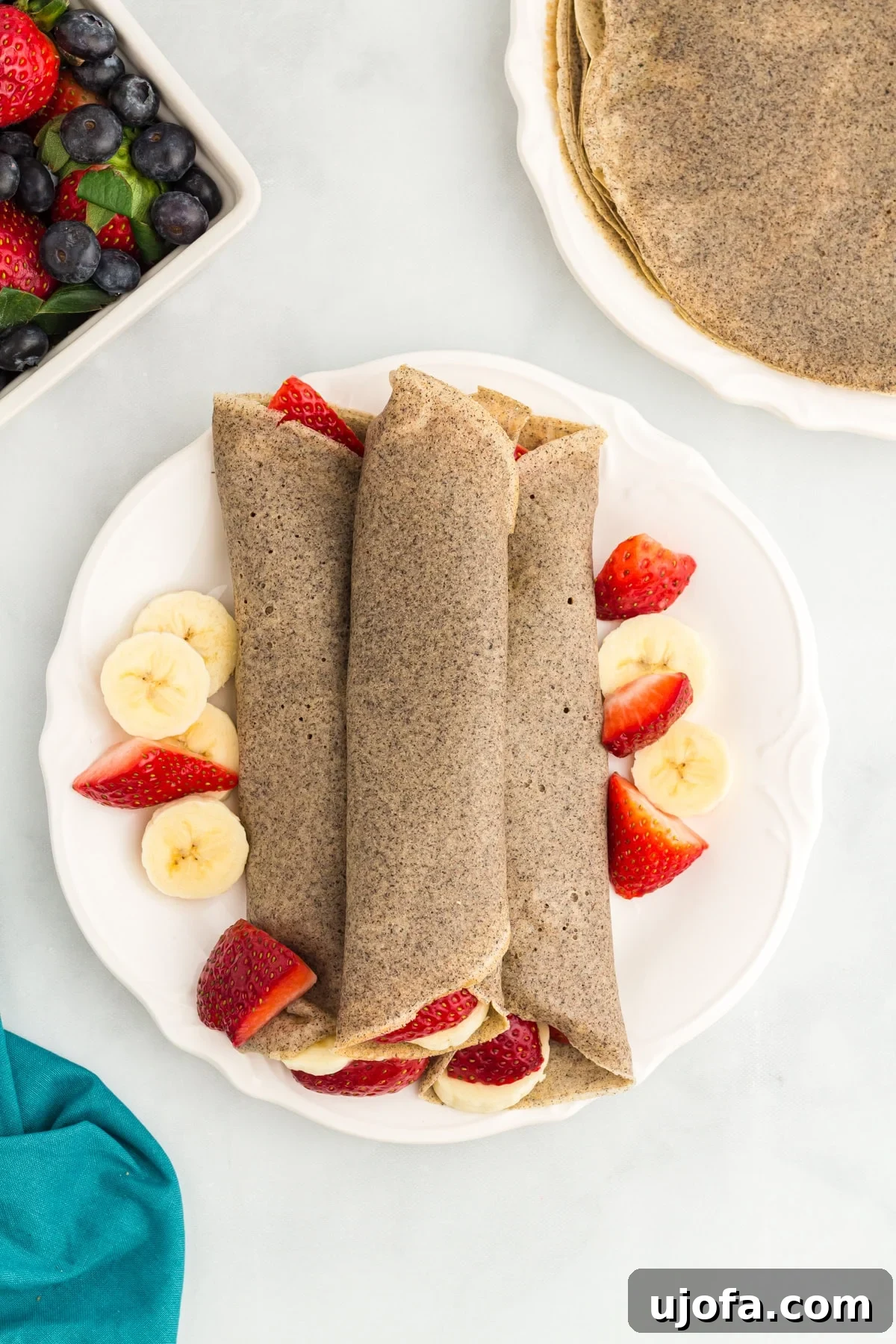 Savory Buckwheat Galettes 2 Three perfectly rolled buckwheat crepes arranged on a pristine white plate, garnished with vibrant fresh banana slices and luscious halved strawberries, ready to be enjoyed.