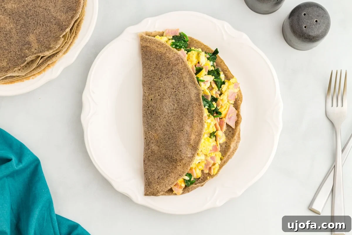 Savory Buckwheat Galettes 11 A savory buckwheat crepe, elegantly folded in half, brimming with a delicious filling of scrambled eggs, savory ham, and tender sautéed spinach, served on a white plate with salt, pepper, and cutlery nearby, presenting a perfect brunch or light meal.