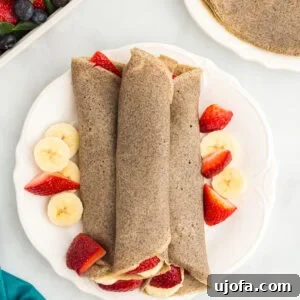 Three rolled buckwheat crepes on a white plate, surrounded by fresh banana slices and halved strawberries.