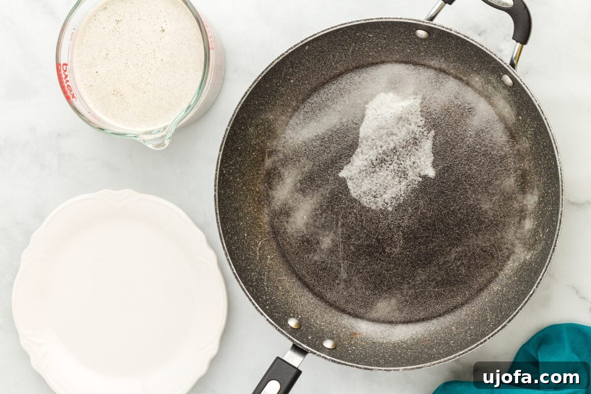 Savory Buckwheat Galettes 6 A non-stick frying pan lightly sprayed with cooking oil sits on a stovetop, next to a measuring cup filled with smooth crepe batter and an empty white plate, ready for the cooking process.