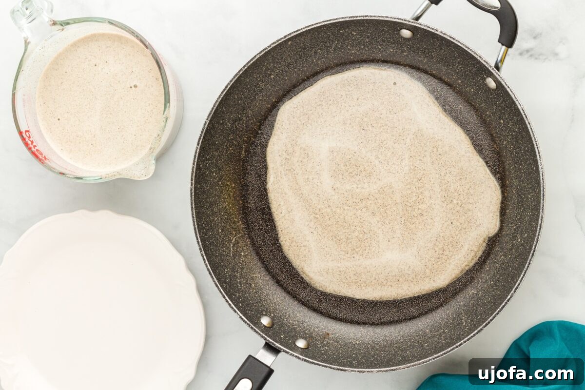 Savory Buckwheat Galettes 7 A thin, circular layer of buckwheat crepe batter cooking in a skillet, gently spreading to the edges. A measuring cup with more batter is visible nearby, indicating the ongoing cooking process.