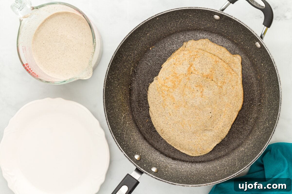 Savory Buckwheat Galettes 8 A buckwheat crepe, perfectly golden-brown on one side, is cooking in a skillet. Next to the pan, a measuring cup holds additional batter, and an empty plate awaits the finished crepes, illustrating the progression of cooking.