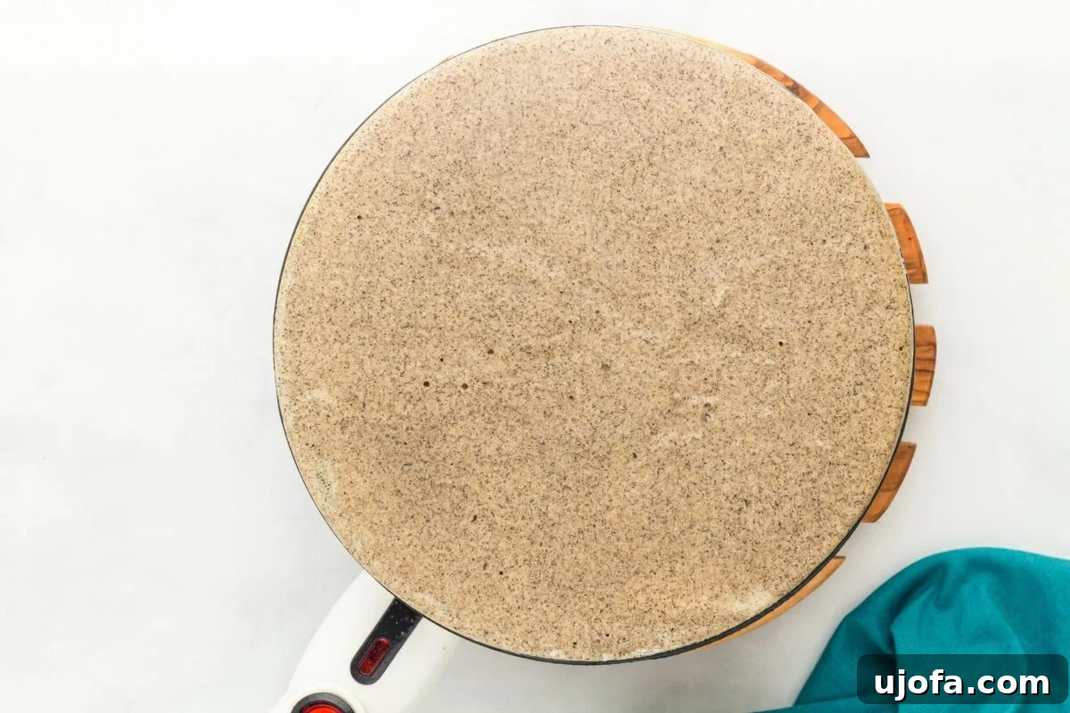 Savory Buckwheat Galettes 9 A thin, circular buckwheat crepe cooking on the flat, heated surface of an electric crepe maker, showing the characteristic lacy texture and even browning.