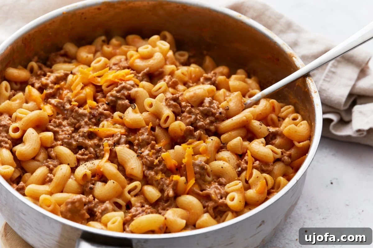 A close-up view of a skillet brimming with cheesy gluten-free Hamburger Helper, its rich, creamy texture highlighted, with a metal spoon resting conveniently on the side, inviting a taste.