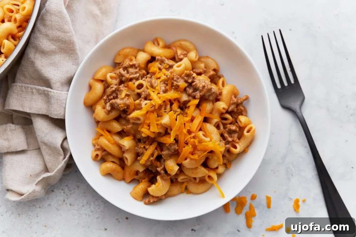 A perfectly portioned serving of gluten-free Hamburger Helper in a pristine white bowl, garnished with melted cheddar, positioned next to the original skillet and a soft beige napkin, ready for a delightful meal.