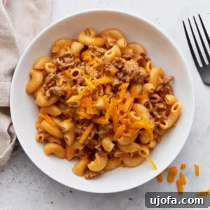 A white bowl filled with creamy gluten-free Hamburger Helper, topped with shredded cheddar cheese, sitting beside a fork on a light surface.